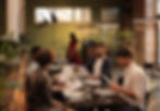 Four people sitting at a table reading menus in a cozy restaurant with green walls and plants. A woman in red walks by. Warm lighting.