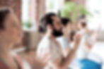 People meditate in a sunlit room with brick walls and greenery. They sit cross-legged, eyes closed, hands together, conveying calmness.