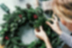 Hands decorate a green Christmas wreath with red berries on a gray surface. Nearby are twine, a pinecone, and a polka-dotted gift.