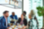 People in a modern restaurant, engaged in conversation. Drinks and plates on the table. Bright lighting, city view outside large windows.