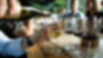 Person pours white wine into a glass on a wooden table with others drinking wine. Blurred faces, casual setting, social and relaxed mood.