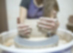 Hands sculpting clay on a spinning pottery wheel. The setting is a workshop, with visible clay and muddy hands. Calm, focused mood.