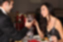 Couple toasting with wine glasses at a candlelit dinner, woman smiling. Red and white decor, rose on table, cozy atmosphere.